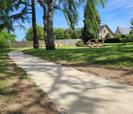 Allée en sable stabilisé sinuant à travers un jardin paysager arboré avec mobilier extérieur