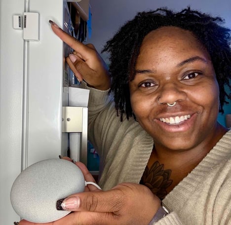 Ashley smiling while holding a smart speaker and pointing to door sensor affixed to refrigerator