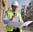 A woman wearing a red safety helmet and yellow earmuffs operates a surveying instrument mounted on a tripod. She is standing in a construction area with a compact excavator in the background. Her casual attire includes a white eyelet blouse and blue jeans, and she is holding a digital level.