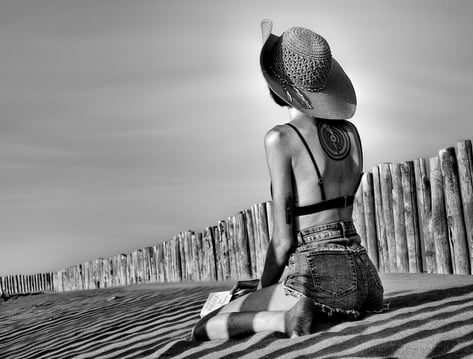 Photo artistique noire et blanc d'une jeune femme en mini short a genoux sur la plage avec un livre et un grand chapeau