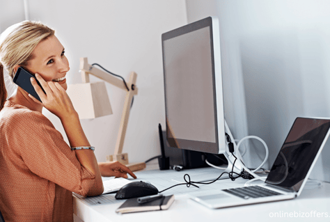 a woman talking on the phone phone while sitting at a desk with desktop and laptop