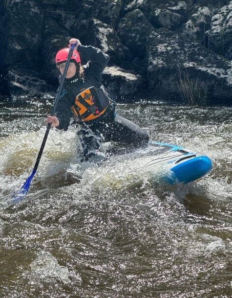 women paddling a Paddleboard over a river wave, wearing helmet, PFD and holding a paddle. 