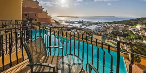 A hotel balcony with two chairs and table, overlooking a swimming pool and Mgarr harbour.