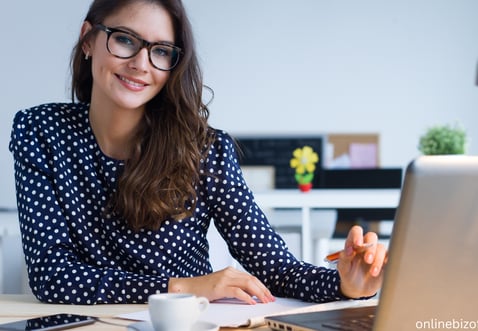 a woman in glasses and a polka dot shirt is smiling and holding a pen with a laptop on the desk