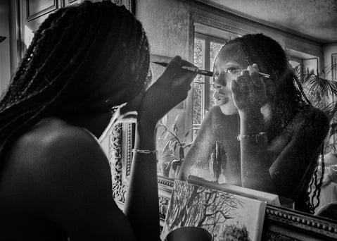 Photo artistique noire et blanc d'une jeune femme noire qui se requi se remaquille dans le miroir d'un intérieur haussmannien