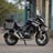 A custom motorcycle with a rugged appearance is parked on a dirt path. The bike is designed with a matte black paint job, featuring a branded logo of 'Cafe Racer'. It is equipped with dual front suspensions, a circular headlamp with grill protection, and handlebars extending outward. The background includes residential buildings and some trees.