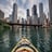 A kayak glides across a calm lake surrounded by lush green forests under a bright blue sky with scattered white clouds. The perspective from the bow of the kayak gives a sense of exploration and adventure.