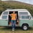 The interior of a camper van features a compact kitchen area with a sink, wooden countertop, and multiple drawers. There are overhead cabinets with warm lighting, along with wood-paneled ceiling and white wall accents. In the background, a cushioned bed is positioned at the rear of the van.