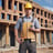 A person wearing a high-visibility safety vest and a beanie stands outdoors, operating a theodolite mounted on a tripod. The background includes the facade of a residential building with balconies.