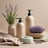A minimalist composition featuring eco-friendly cleaning products arranged on a textured beige fabric. There's a bottle with a pump dispenser, a small packet, and two paper bags with lavender and mint green labels. The background is softly lit, creating a calm atmosphere.