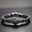 A collection of intricately designed metallic jewelry pieces in various shapes, featuring floral and leaf motifs. The pieces are adorned with small, round, deep green beads scattered on a textured dark surface.