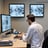 Two people in lab coats collaborate in an office setting. One is seated and pointing at a computer screen displaying medical images, while the other stands nearby holding a tablet. Large windows reveal a view of greenery outside, creating a bright and professional environment.