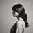 A ballet dancer is captured from behind, standing on pointe. She wears a black leotard with lace detailing on the back and a flowing white tulle skirt. Her hair is styled in a neat bun, and she is poised gracefully in a minimalist, light-filled space.