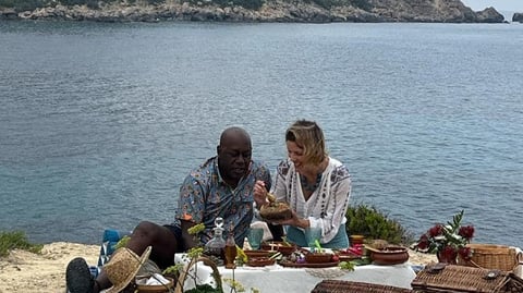 A man and woman sit on the floor, behind a low table of food, behind them is the sea.