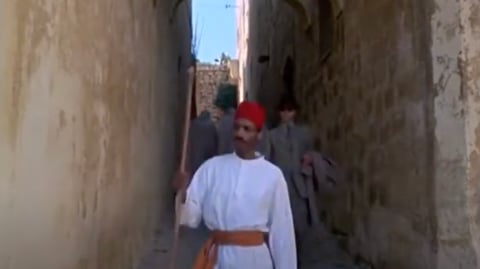 A man in a white robe and a fez walks down a narrow alley.