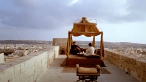 Two men sit under an elaborate canopy, overlooking a city.