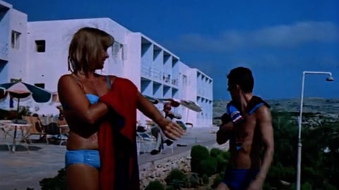 A man and a woman in bathing suits stand in front of a hotel.