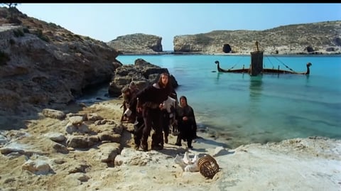 Group of men standing on a beach, in front of a half sunken ship.