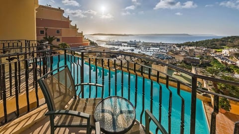 A hotel balcony with two chairs and table, overlooking a swimming pool and Mgarr harbour.