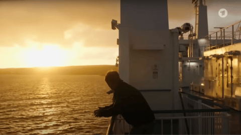 A man leans against the railing of a ship as the sun sets in the distance.