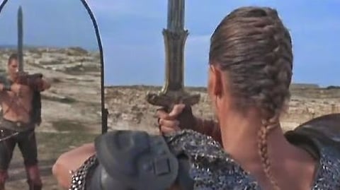 A man in armour and chain mail faces himself in a mirror, atop a cliff.