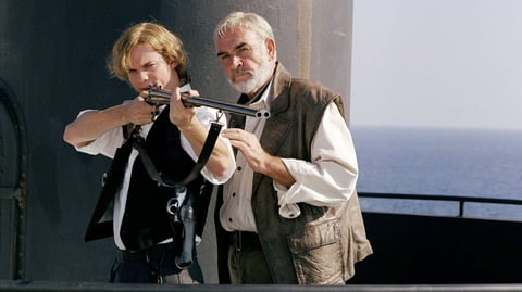 A man holding a gun in front of a submarine conning tower, while another man tutors him.