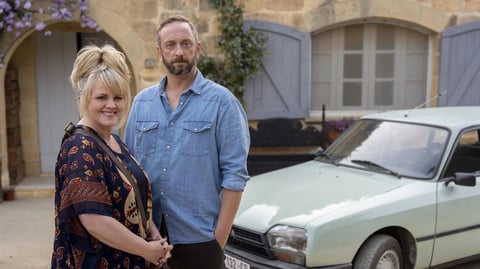 Jean Whiite and Dom from The Madame Blanc Mysteries, standing in front of a tradtional cottage next to a car. 