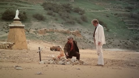 Two men stand on a beach by a grave.