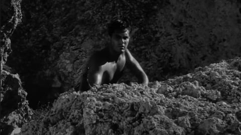 A bare-chested man hides behind a rock.