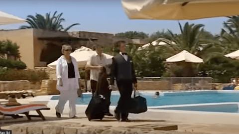 A man and woman are being escorted past a hotel pool a porter, who is carrying their luggage.