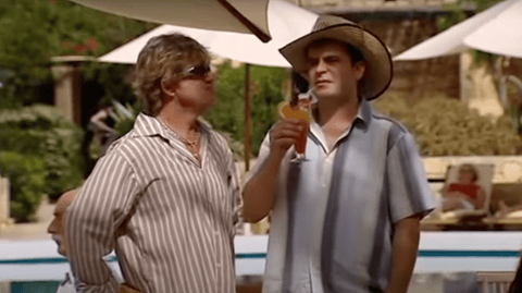 Two men stand underneath a sun parasol  in front of a hotel pool.