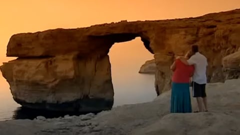 A man and woman stand looking at the sunset through the Azure Window. He has his arm around her shoulder.