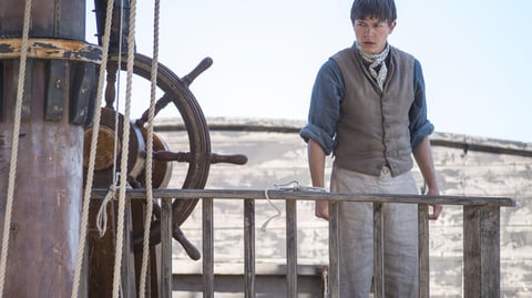 A teenage boy stands on the deck of a sailing ship.