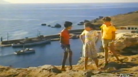 Three children standing on a cliff overlooking a harbour. 