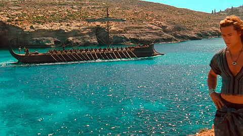 A man dressed as an ancient Greek stands on a cliff in front of a body or water with a galley.