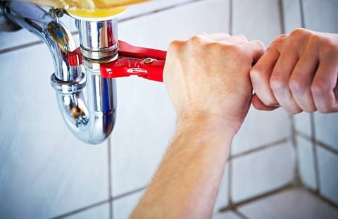 man using a red mini pipe wrench to turn a pipe under the sink