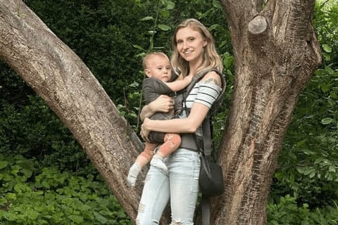 Mother holding baby in a carrier by a tree, bonding in nature.
