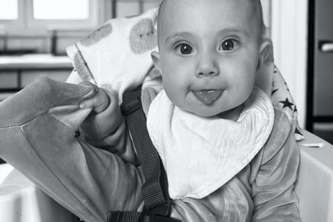 Black and white photo of smiling baby in high chair with tongue out, showing natural baby joy, innocence, and purity.