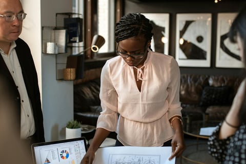 One man and two women standing at a table discussing business blue print schematics with a laptop displaying charts