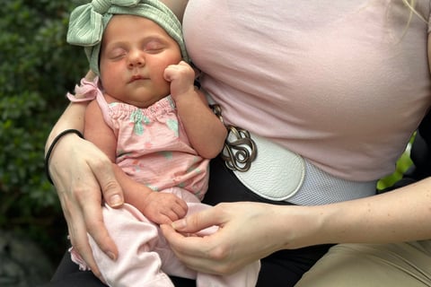 A mother in a pink shirt holding her sleeping baby in a green bow resting with hand on cheek.