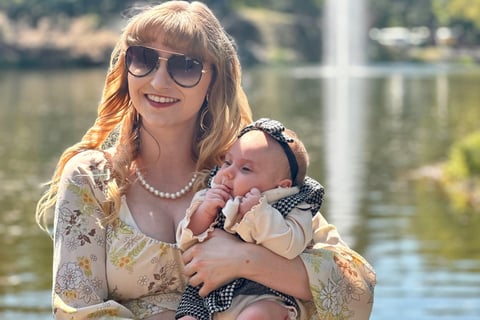 A mother cradling her newborn daughter in front of a lake, with a fountain softly spraying in the background.