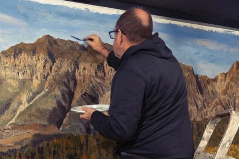 Martin Osner painting a large mountain landscape on canvas using acrylics and a palette, painting on a ladder