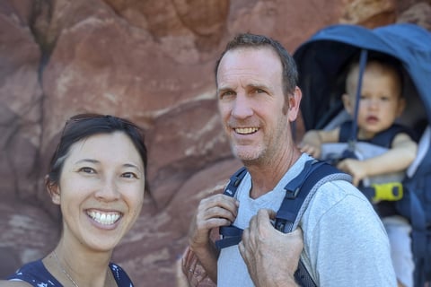 Smiling couple hiking with their baby in a carrier against red rock cliffs in Colorado.