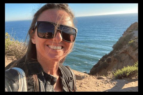 Michael Alexa Pitts on a rocky Pacific coastline with ocean behind, smiling, with black sunglasses & strands of hair in face