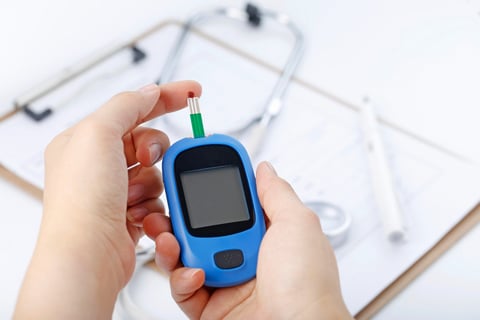 Hand holding a blood glucose meter measuring blood sugar, the background is a stethoscope and chart
