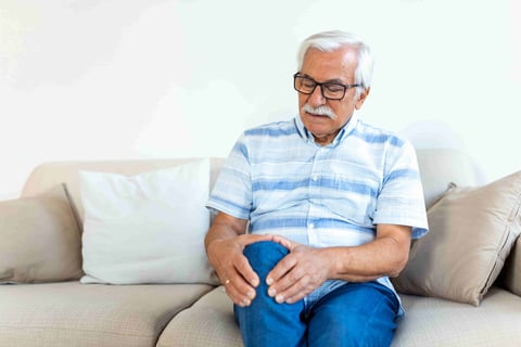 Elderly man sitting on a sofa at home and touching his painful knee.