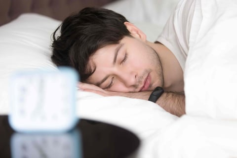 Young man sleeping next to alarm clock wearing smart wristband.