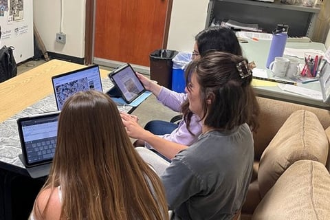 Three students sitting at a table, each with a computer or tablet in front of them reviewing video