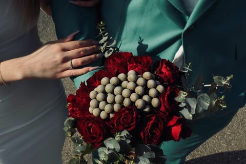 Buchet cu trandafiri roșii, craspedia și eucalipt, ținut de un cuplu.