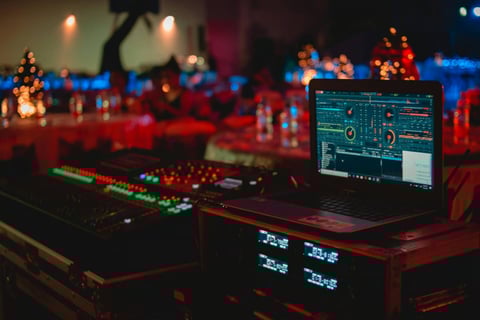 A laptop running DJ software sits on a table with audio equipment and mixers, with blurred decorative lights in the backgroun